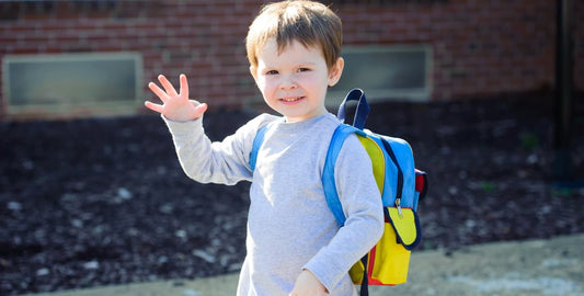 Niño pequeño con mochila de colores saludando en su primer día de colegio